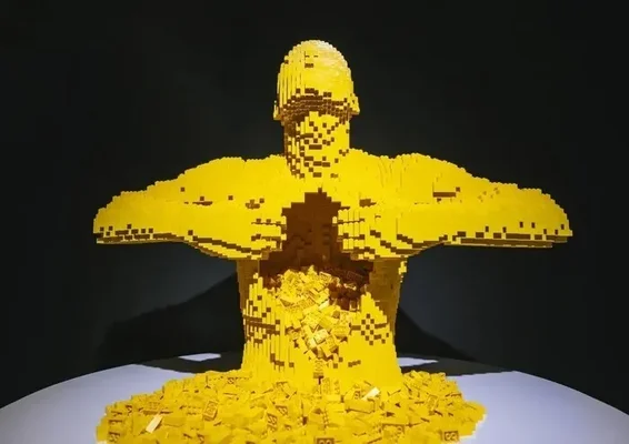 Sculpture LEGO jaune représentant un torse humain aux bras tendus qui semble fondre, avec des briques éparpillées sur une surface blanche, à l'exposition The Art of the Brick à Nantes.