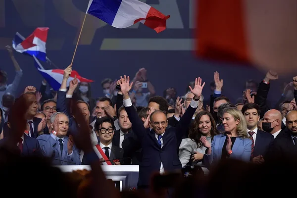 Éric Zemmour, en costume sombre, les bras levés devant des partisans agitant des drapeaux français lors de son premier meeting à Villepinte en décembre 2021.