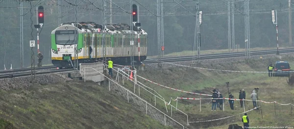 Un train de voyageurs immobilisé sur la voie ferrée près de signaux rouges, des agents en gilets haute visibilité et un groupe de personnes derrière une bande de police sur le talus après l'explosion