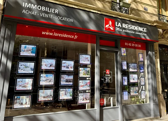 Devanture de l'agence immobilière La Résidence à Paris 17e, façade grise et rouge avec de grandes vitrines où sont exposées de nombreuses annonces immobilières.