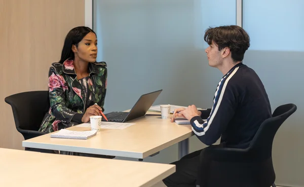 Une femme et un homme assis face à face autour d'une table claire en bureau, avec un ordinateur portable, des documents, des carnets et des tasses à café, lors d'un entretien ou d'une réunion.
