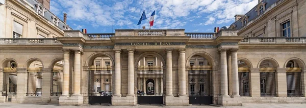 Façade en pierre du Conseil d’État à Paris, avec arches, grilles en fer, colonnes classiques, inscription « CONSEIL D’ETAT » et drapeaux français et européen sous un ciel bleu.