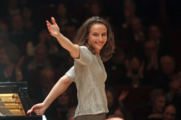 Hélène Grimaud, cheveux bruns ondulés et haut argenté scintillant, debout devant un piano à queue sur scène, salue d'un bras en souriant tandis que le public applaudit en arrière-plan.