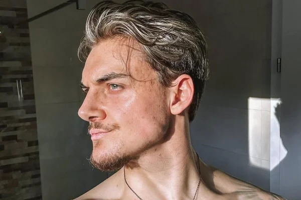 Un homme torse nu, cheveux bicolores coiffés, moustache et bouc, portant un collier fin et un tatouage à l'épaule, se tient dans une salle de bains et regarde vers la gauche.