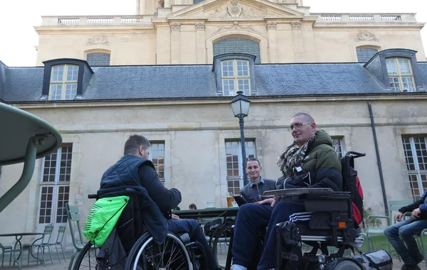 Dans une cour ensoleillée, deux hommes en fauteuil roulant, dont un en fauteuil électrique, discutent avec un homme attablé devant un bâtiment historique en pierre de l'hôpital des « gueules cassées » à Paris.