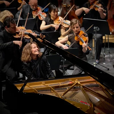 Hélène Grimaud, assise à un piano à queue en concert, sourit vers des violonistes de l'orchestre derrière elle, au Festspielhaus de Baden-Baden.