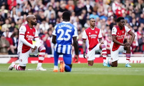Quatre joueurs de football, dont trois en maillot rouge et blanc d'Arsenal, posent un genou à terre sur la pelouse avant un match, avec un public flou en arrière-plan.