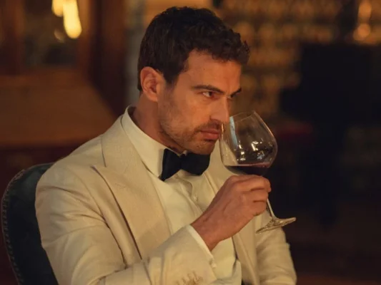 Theo James, en smoking blanc et nœud papillon noir, assis dans un décor feutré, hume un verre de vin rouge dans la bande-annonce de The Gentlemen saison 2.