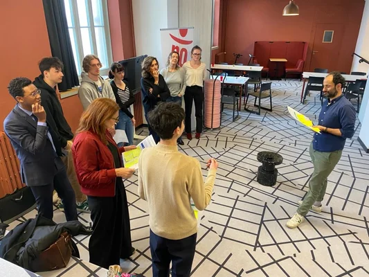 Groupe d’une dizaine de personnes réunies dans une salle au sol noir et blanc à Sciences Po Aix, écoutant un homme en chemise bleue tenant des documents lors de la convention sur l’IA générative.