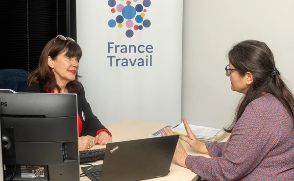Deux femmes assises face à face à un bureau lors d'un entretien avec une conseillère France Travail, dans un bureau où le logo France Travail est visible à l'arrière-plan.