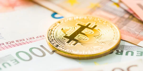 Une pièce physique dorée de Bitcoin posée sur un billet de 100 euros, entourée d'autres billets en euros