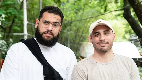 Deux hommes debout en extérieur devant un feuillage vert : à gauche, un homme barbu portant des lunettes et une chemise blanche de type thobe avec un vêtement noir sur l'épaule ; à droite, un homme souriant en t-shirt beige et casquette claire, dans l'émission « Follow The Leader » consacrée aux pas