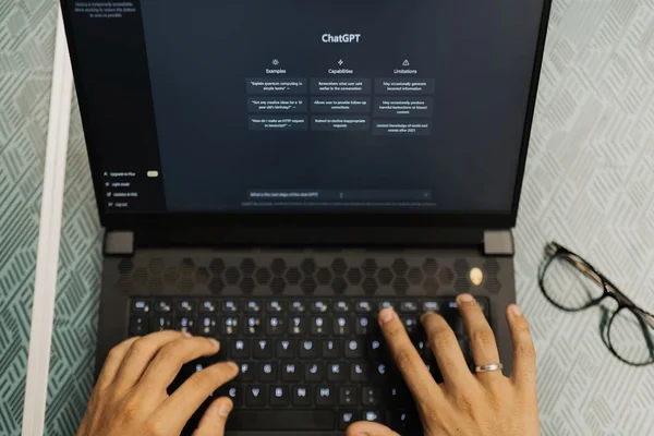 Vue en plongée de mains tapant sur le clavier d’un ordinateur portable affichant l’interface ChatGPT, avec une paire de lunettes posée à côté.