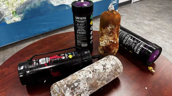 Quatre bouteilles de protoxyde d'azote posées sur une table en bois : deux bouteilles noires à chapeau violet portant une étiquette de danger ONU 1070, à côté de deux bouteilles fortement corrodées et couvertes de rouille.