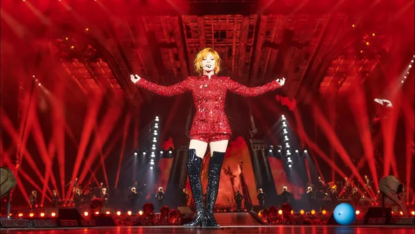 Mylène Farmer sur scène lors de Nevermore, en tenue rouge à sequins et bottes noires montantes, bras écartés sous des lumières rouges, entourée de danseurs en silhouette.