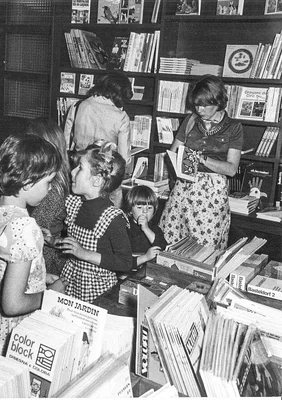 Photographie en noir et blanc d'enfants et d'adultes feuilletant des albums empilés sur une table, dans une salle aux hautes étagères entièrement garnies de livres, chez L'École des loisirs.