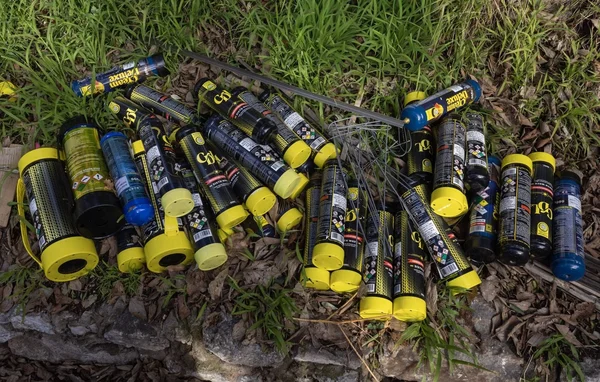 Bonbonnes cylindriques métalliques, dont plusieurs modèles à capuchon jaune, jonchées sur un sol herbeux et terreux au pied d'une bordure en pierre.