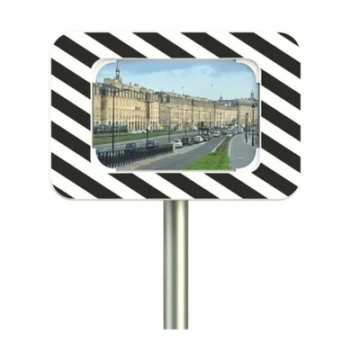 Miroir de sécurité routier rectangulaire à cadre noir et blanc rayé en diagonale, monté sur un poteau métallique, avec au centre un visuel d'une rue urbaine courbe bordée de bâtiments en pierre et de voitures garées.
