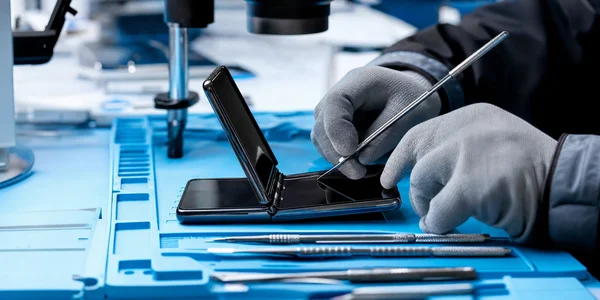 Mains gantées d'un technicien réparant l'écran d'un Samsung Galaxy Z Flip 2 ouvert sur un tapis antistatique bleu, avec tournevis de précision et pinces au premier plan.