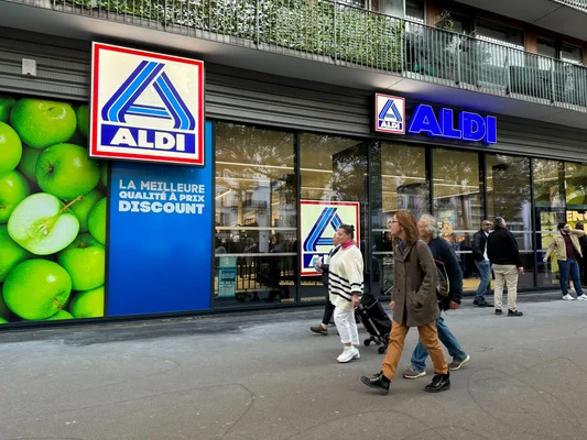 Devanture d'un magasin Aldi à Paris, avec logo, signalétique discount et grande pomme verte, des piétons marchant sur le trottoir devant l'entrée.