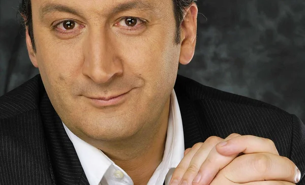 Portrait studio en gros plan de Sylvain Bourmeau, cheveux courts bruns, en veste sombre à fines rayures sur chemise blanche ouverte, souriant légèrement, les mains jointes devant lui sur fond gris anthracite moucheté