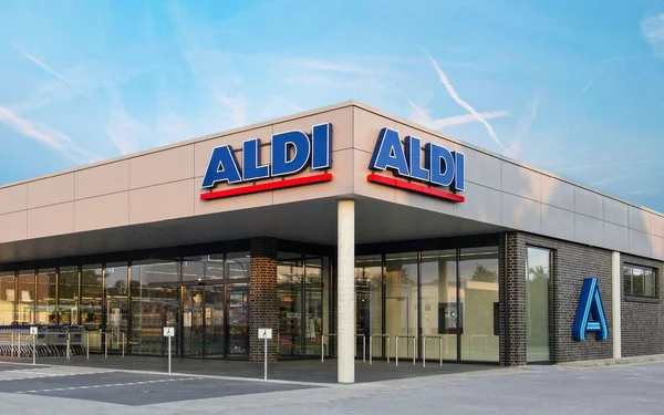 Façade moderne d'un magasin Aldi de jour, avec grandes vitrines, enseignes ALDI, logo bleu sur un mur de briques et parking devant sous un ciel bleu.