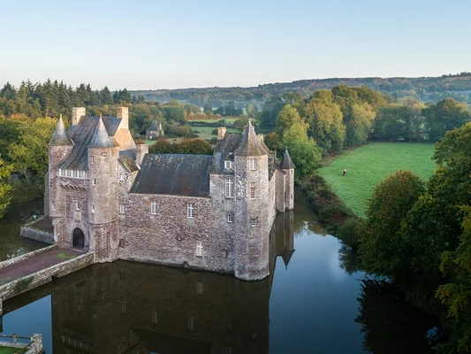 Vue aérienne du château de Trécesson à Campénéac, forteresse médiévale en pierre entourée de douves, avec un pont d'accès, des arbres d'automne, des champs verts et des collines.