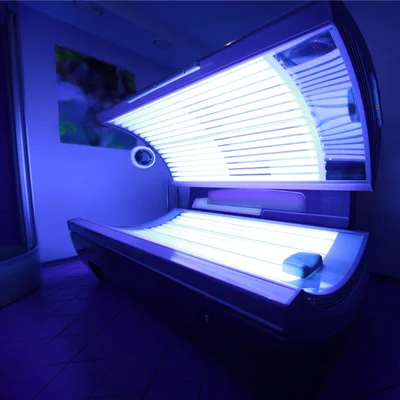 Lit de bronzage horizontal ouvert, tubes ultraviolets allumés diffusant une lumière bleutée sur les parois carrelées d'une cabine