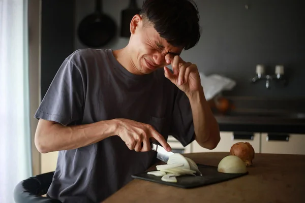 Un homme en t-shirt gris coupant un oignon sur le plan de travail de sa cuisine, grimaçant et portant la main à son œil à cause des vapeurs irritantes