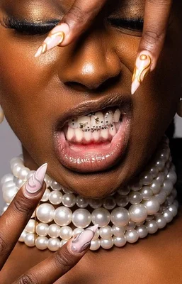 Gros plan éditorial sur une personne portant des bijoux dentaires dorés et des tooth gems, avec un collier de perles et des ongles manucurés ornés d'accents dorés.