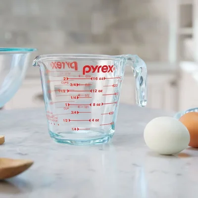 La tasse à mesurer Pyrex de 2 tasses, en verre transparent avec graduations rouges, posée sur un comptoir en marbre avec deux œufs, une cuillère en bois et un bol en verre.