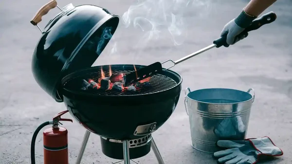 Une personne portant un gant résistant à la chaleur utilise un outil à long manche pour remuer les braises d'un barbecue à charbon noir, avec un extincteur et un seau d'eau à proximité.