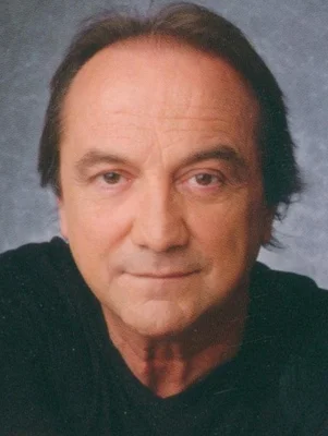 Portrait de Claude Dubois, chanteur québécois, cheveux bruns clairsemés et haut noir à col en V, posant devant un fond gris moucheté.