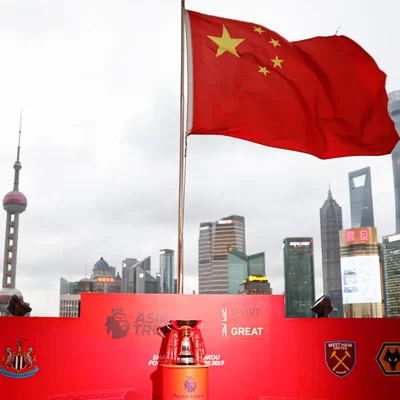 Drapeau chinois flottant en premier plan devant le skyline de Shanghai avec la Tour de la Perle d'Orient, et un écran rouge présentant le trophée Premier League Asia et les logos des clubs.
