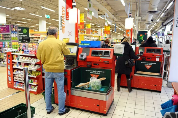 Un homme en veste jaune et jean bleu utilise une caisse automatique rouge dans un supermarché ; une autre cliente est à une caisse voisine, avec rayonnages et signalétique en arrière-plan.