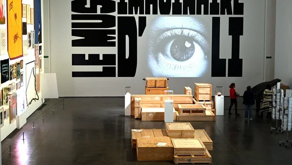 Vue de l'exposition du Musée imaginaire d'Oli au Grand Palais : une grande installation murale avec des lettrages noirs encadrant la projection d'un oeil humain, des caisses de transport en bois disposées sur le sol poli et quelques visiteurs qui déambulent