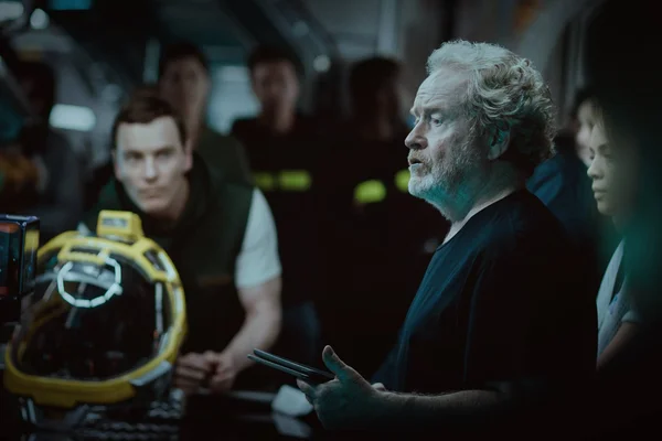 Ridley Scott, cheveux gris ondulés et barbe grise, vêtu de noir, tablette à la main sur un plateau sombre, avec un casque jaune au premier plan et des membres d'équipe en arrière-plan.