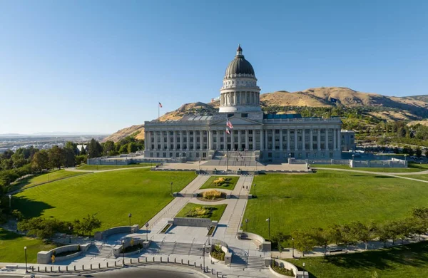 Le Capitole de l'État de l'Utah, à Salt Lake City : un vaste bâtiment gouvernemental surmonté d'un dôme, entouré d'une pelouse verte, avec des collines arides en arrière-plan sous un ciel bleu dégagé.
