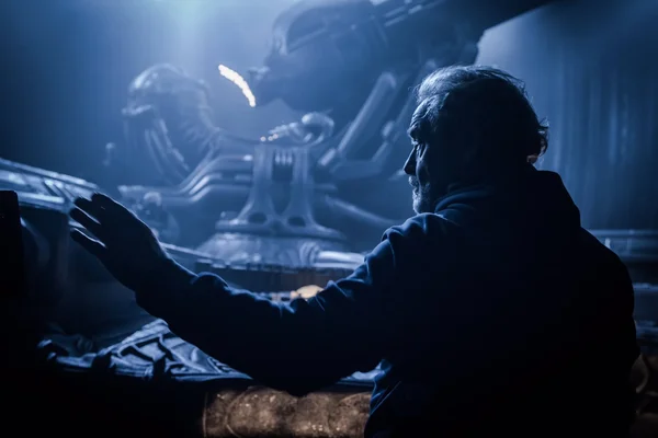 Un homme aux cheveux gris, vu de profil dans une lumière bleue, tend la main vers une créature extraterrestre allongée dans une chambre de science-fiction sombre.