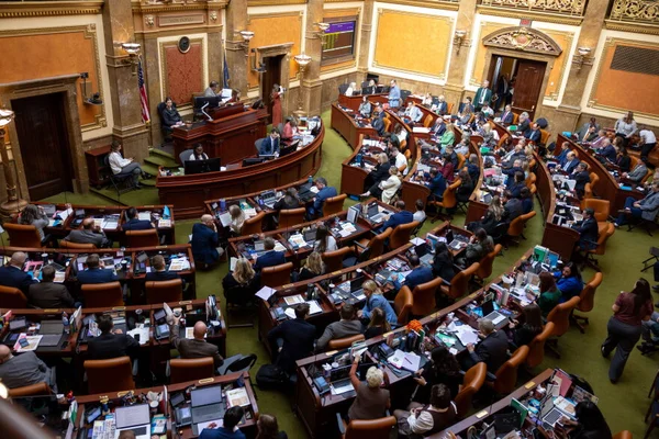 Vue plongeante sur l'hémicycle de la Chambre des représentants de l'Utah en pleine session : les élus sont assis à des rangées courbes de pupitres couverts d'ordinateurs portables et de documents, face au pupitre de présidence, dans un décor de boiseries et de dorures.