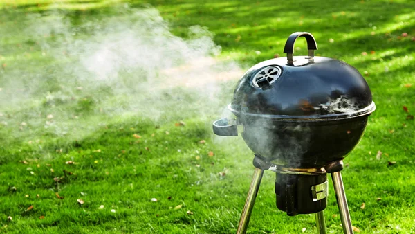 Un barbecue à charbon noir de type boule est installé sur une pelouse et laisse s'échapper une fumée blanche par ses évents et son couvercle.