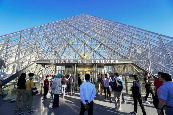 Photographie de la pyramide en verre d'entrée du Musée du Louvre à Paris, avec une foule de visiteurs devant sous un ciel bleu