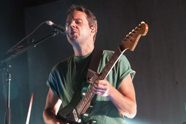 Anthony Gonzalez, de M83, chante au micro tout en jouant d’une guitare électrique vert foncé lors d’un concert à The Anthem, en t-shirt vert et oreillette.