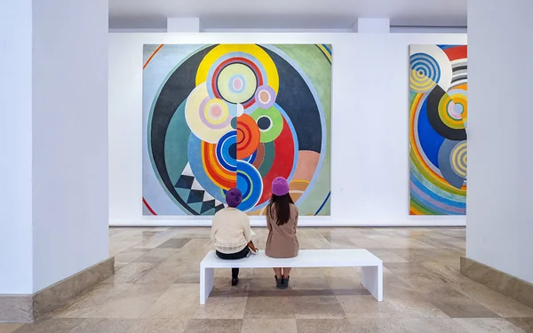 Deux personnes portant des chapeaux violets assises sur un banc blanc dans une galerie d'art, observant de grands tableaux abstraits colorés