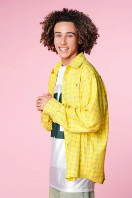 Jeune homme aux cheveux bouclés portant une chemise à carreaux jaune sur un t-shirt blanc, souriant face à l'objectif sur fond rose, candidat de la Star Academy 2024.