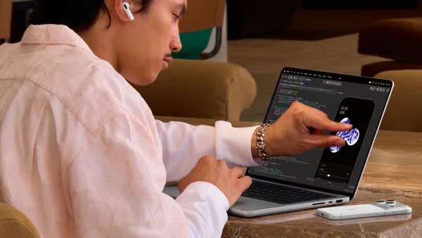 Un jeune homme en chemise rose et écouteurs blancs utilise un MacBook Pro à écran tactile affichant du code et une interface d'application mobile, avec un smartphone posé à côté.