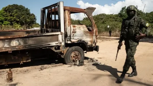 Photographie d'un camion-citerne brûlé et rouillé stationné sur une route de terre, un soldat en tenue tactique, arme à l'épaule, passant à côté