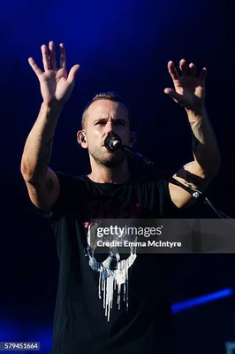 Anthony Gonzalez, fondateur de M83, sur scène, les mains levées, en t-shirt noir à motif graphique, baigné d’une lumière bleue.