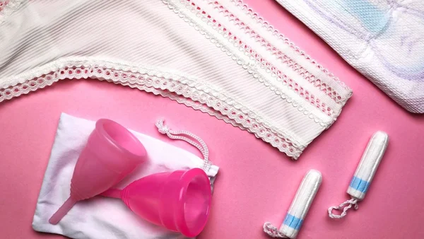 Divers produits d'hygiène menstruelle, y compris des coupes menstruelles roses, des tampons, une serviette et des sous-vêtements, sur un fond rose.