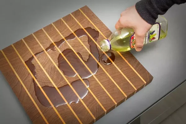 Main versant de l'huile depuis une bouteille sur une planche à découper en bois à rayures claires, où un liquide sombre s'est déjà accumulé